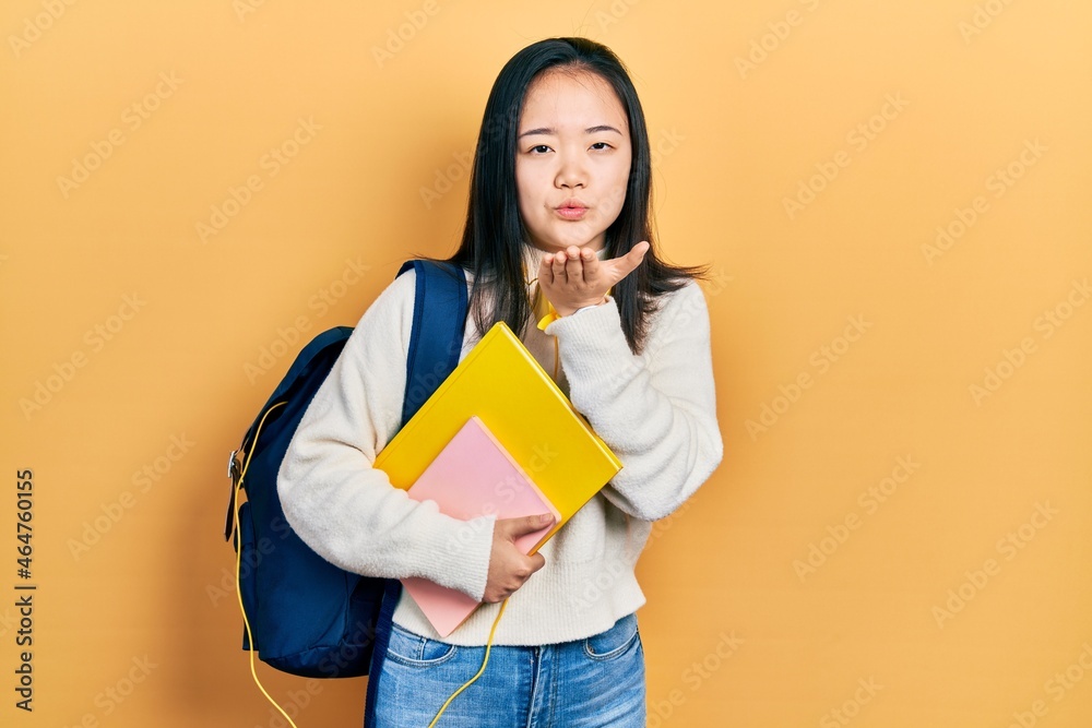 Young chinese girl holding student backpack and books looking at the camera blowing a kiss with hand on air being lovely and sexy. love expression. Young chinese girl holding student backpack and books looking at the camera blowing a kiss with hand on air being lovely and sexy. love expression.