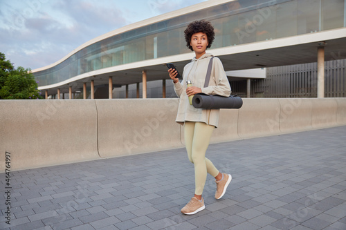 Sporty motivated woman in activewear carries karemat holds bottle with water and smartphone checks receieved notifications whilehaving workout walks outdoors has regular training. Healthy lifestyle photo