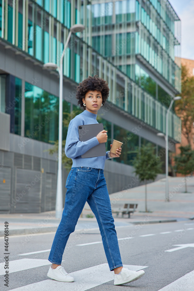 Outdoor shot of fashionable student carries modern tablet and takeaway coffee walks on crossroad looks away poses in downtown uses application returns from studying. Modern lifestyle concept