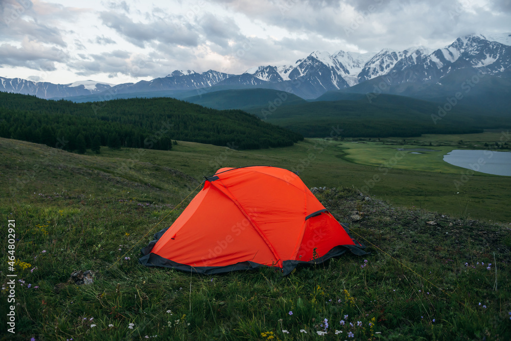 Atmospheric alpine landscape with orange tent on background of lake and big snowy mountains in overcast weather. Awesome green scenery with orange tent with view of great mountains under cloudy sky.