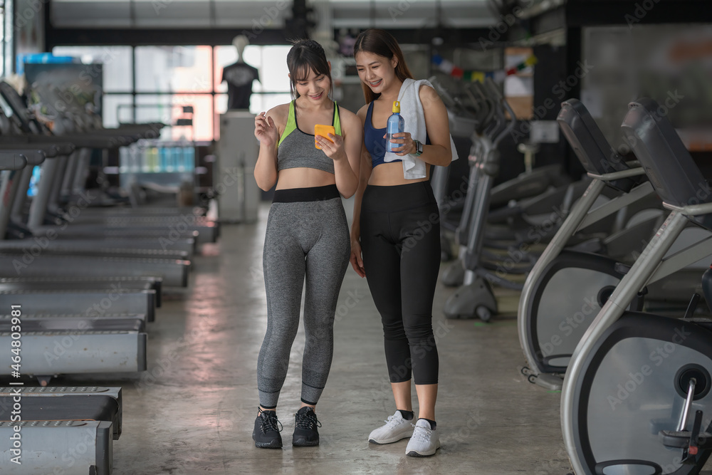 Fototapeta premium A smiling fit lady is showing her female friend something on phone. Both girls are resting after training together in the sports center.