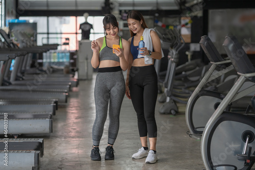 A smiling fit lady is showing her female friend something on phone. Both girls are resting after training together in the sports center.