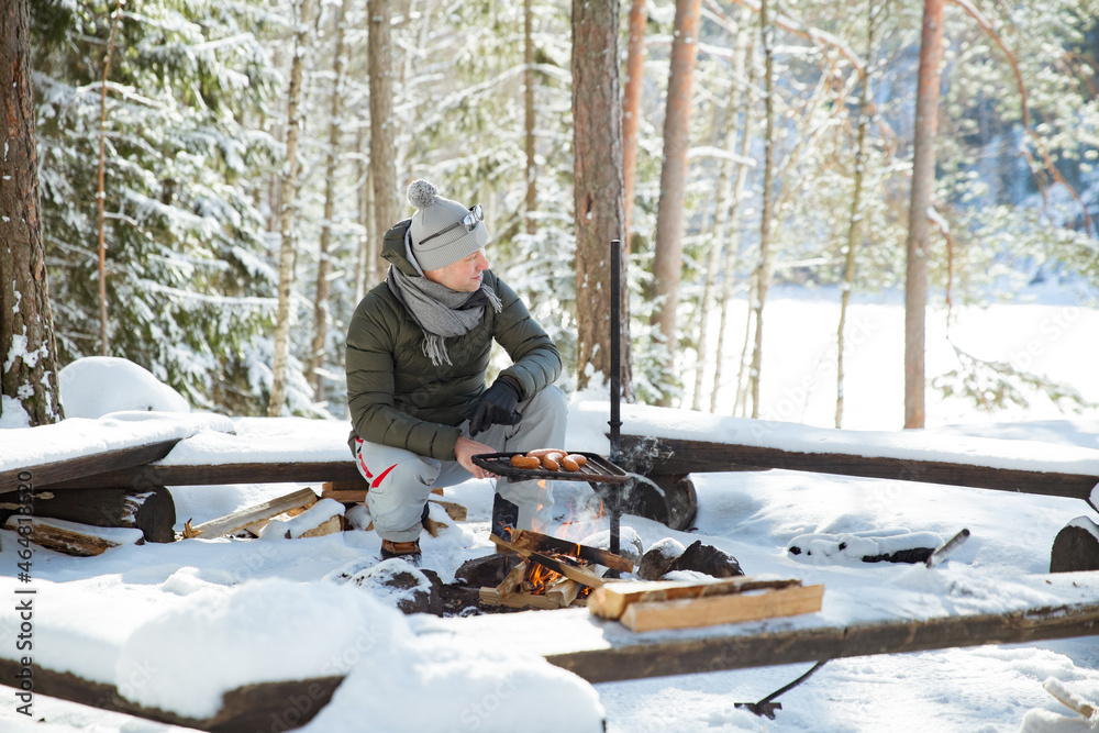 Man roasting sausages on campfire in forest by the lake, making a fire, grilling. Happy tourist exploring Finland. Beautiful sunny winter landscape, wood covered with snow. Man roasting sausages on campfire in forest by the lake, making a fire, grilling. Happy tourist exploring Finland. Beautiful sunny winter landscape, wood covered with snow.