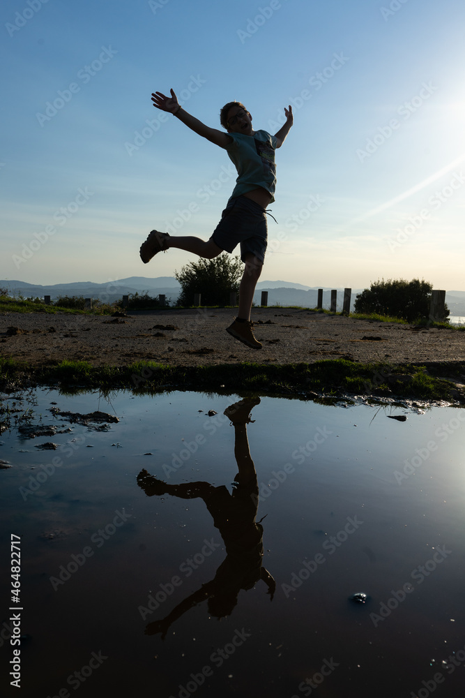 silhouette of a child jumping amused in front of a puddle of water. on the puddle you can see his reflection.