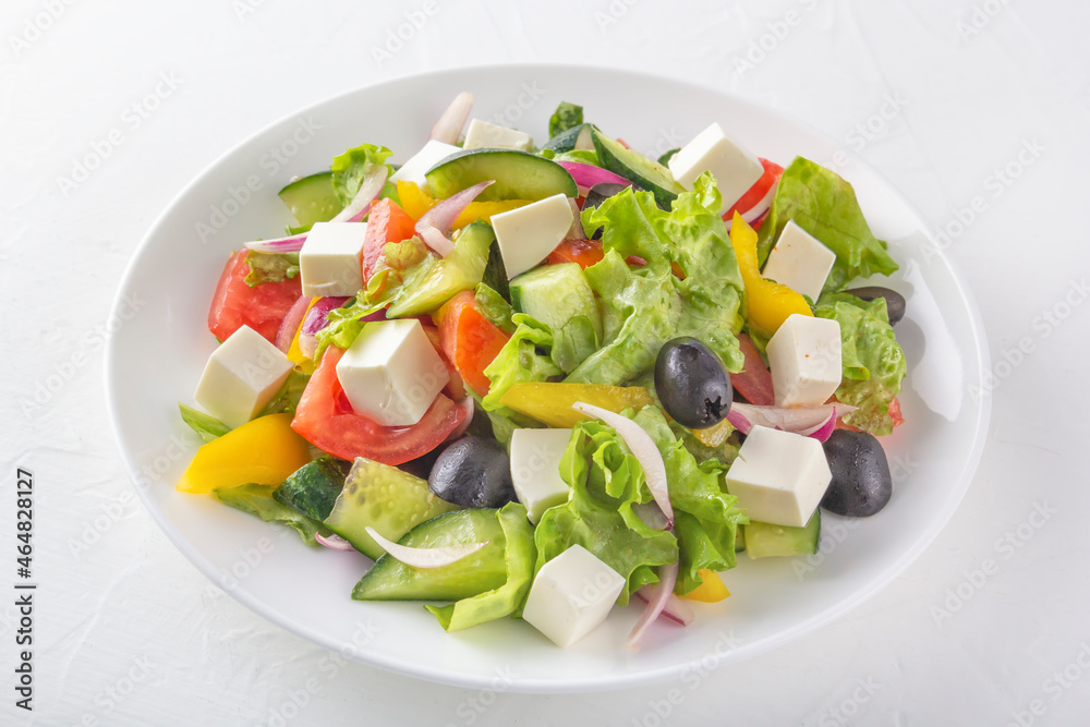 Greek salad of fresh cucumber, tomatoes, bell peppers, lettuce, red onions, feta cheese and olives with olive oil in a white plate on a light background. Healthy eating. Close up