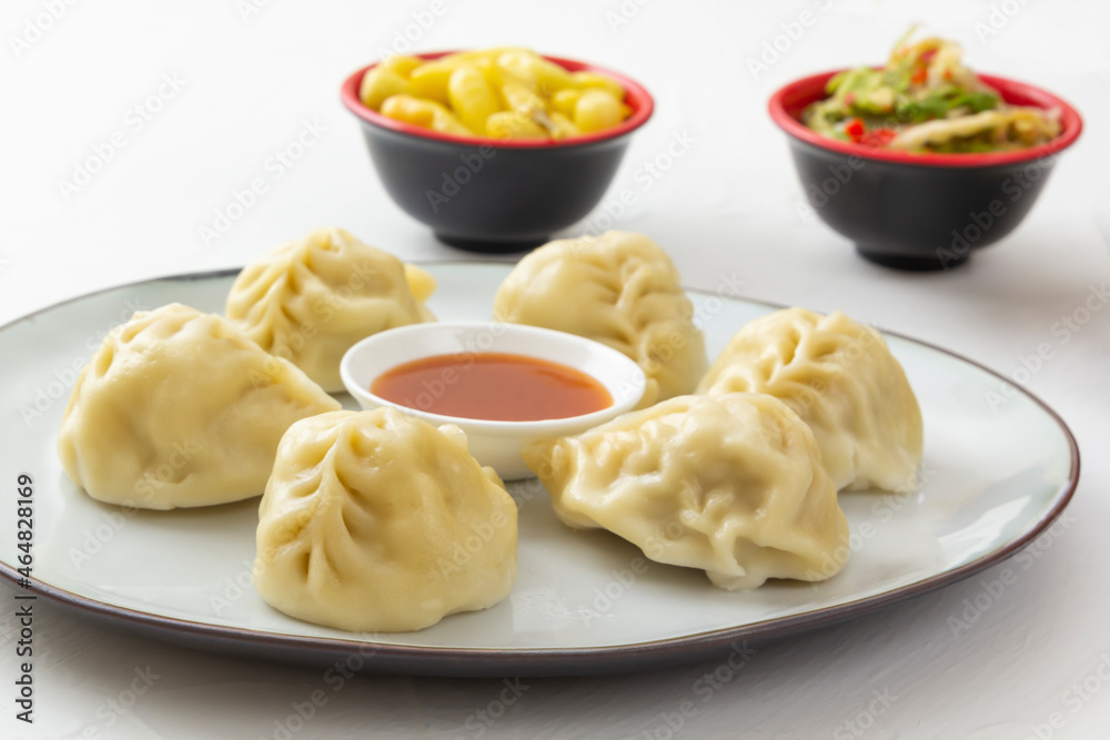 Boiled manti, dumplings or wonton dough stuffed with minced meat in a plate with sauce on a white table against a background of pickled mushrooms and peppers. Homemade food. Close-up Boiled manti, dumplings or wonton dough stuffed with minced meat in a plate with sauce on a white table against a background of pickled mushrooms and peppers. Homemade food. Close-up