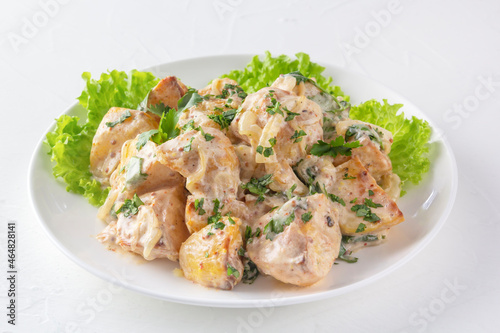 Baked potatoes in sour cream sauce with herbs are placed in a white plate on lettuce leaves. Close-up