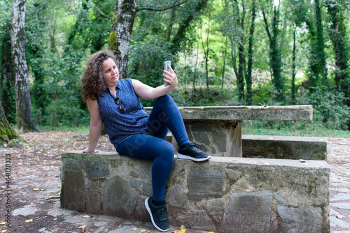 Woman with slightly curly hair sitting on a picnic bench taking a selfie with her smartphone.