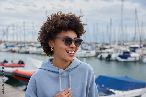 Happy curly haired woman wears casual hoodie walks in marina during spare time enjoys beautiful biews poses in port against ships and yachts spends vacation at seaside town strolls along coast #464829391