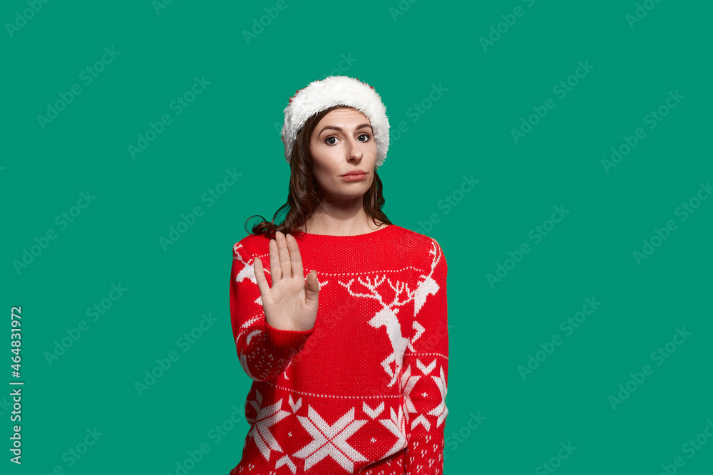 Serious woman with dark hair shows stop sign with hand, refuses of a gift she does not like, stands her ground, wears red ugly sweater with deer and santa hat on green background. Christmas concept.