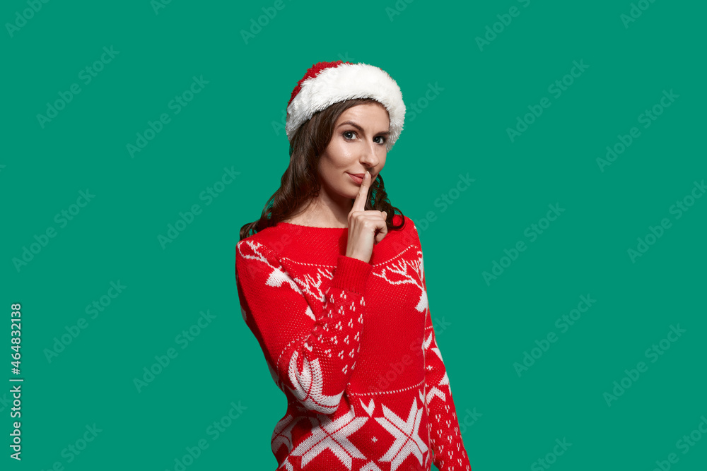 Playful brunette presses index finger to lips, asking to keep quiet and wait, makes surprise for her boyfriend, wears red ugly sweater with deer and santa hat on green background. Christmas concept. Playful brunette presses index finger to lips, asking to keep quiet and wait, makes surprise for her boyfriend, wears red ugly sweater with deer and santa hat on green background. Christmas concept.