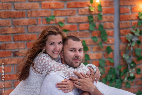 Loving couple millennials are hugging on white bed against background of brick wall with green ivy
