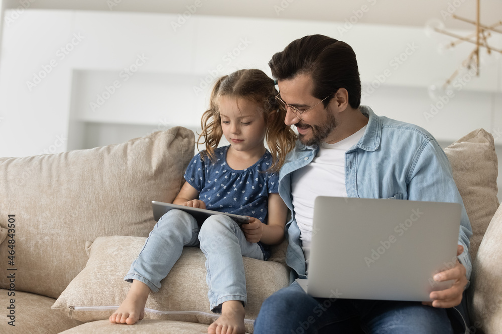 Bonding happy young father and little cute kid daughter holding different gadgets, small child girl playing on digital tablet while smiling daddy using computer, modern tech addiction concept. Bonding happy young father and little cute kid daughter holding different gadgets, small child girl playing on digital tablet while smiling daddy using computer, modern tech addiction concept.