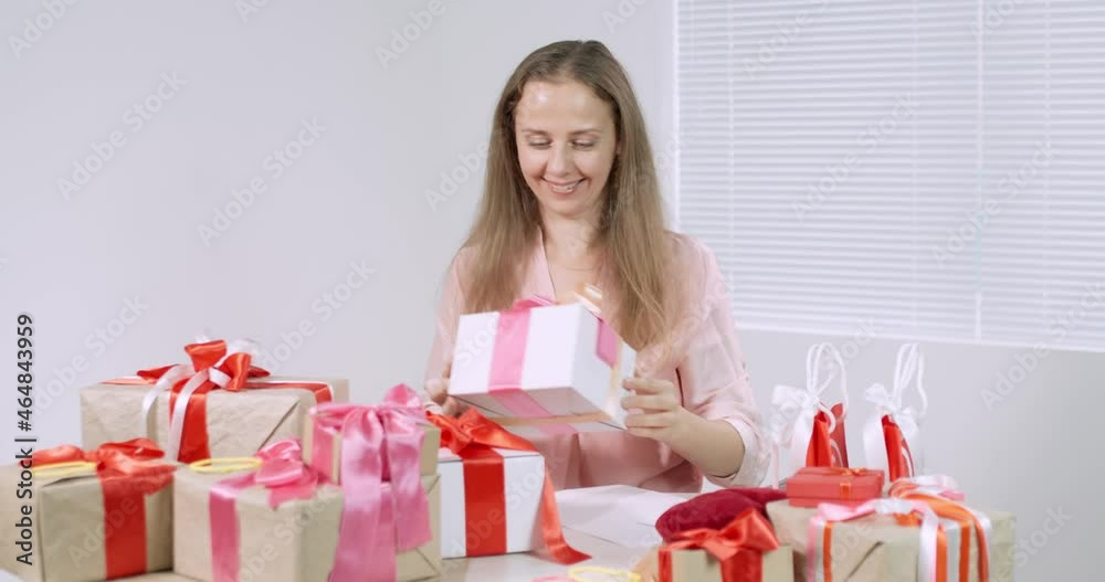 a woman sits at a table with a bunch of gifts, chooses one box and unfastens a bow, looks at what is in the box, rejoices at the gift a woman sits at a table with a bunch of gifts, chooses one box and unfastens a bow, looks at what is in the box, rejoices at the gift