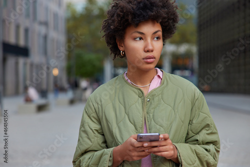 Horizontal shot of thoughtful woman wears jacket holds mobile phone looks away on promenade checks notification poses outside against blurred background stands at street shares publication in blog