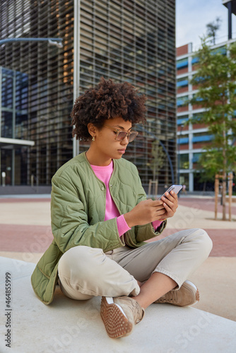 Fashionable female blogger sits crossed legs against urban setting watches video on website via smartphone connected to wireless internet chats in social network wears sunglasses stylish clothes