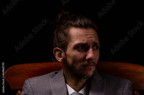 A young businessman in a smart gray suit sitting in a wooden arm chair with moody and atmospheric lighting