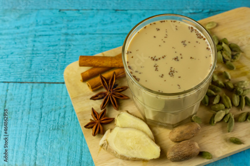 Glass of Masala chai tea with ginger, cardamom, cinnamon, nutmeg, and anise on cutting board on blue wooden background. Masala chai popular traditional India hot drink made of milk, tea and spices.