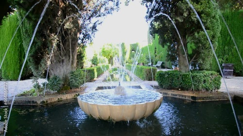 Outdoor fountain made of marble with water flowing in the beautiful gardens of the famous Alhambra, Granada, Spain. Green plants at both sides.