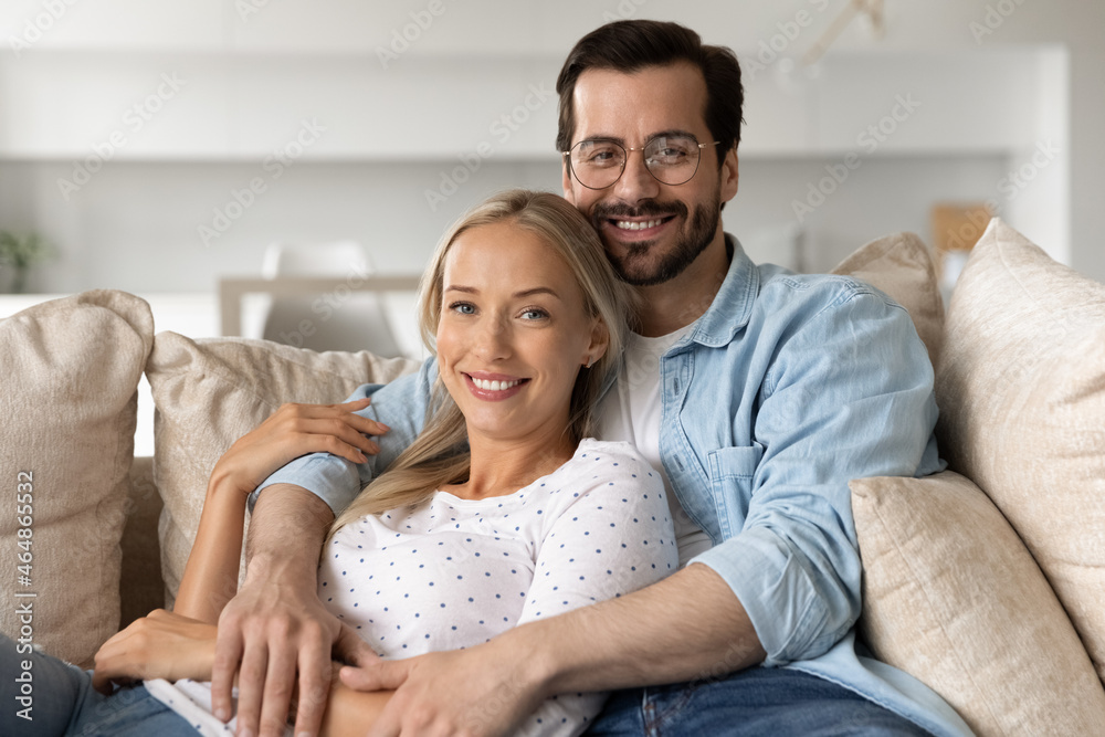 Portrait of smiling beautiful bonding young family couple resting on cozy sofa. Happy affectionate millennial man cuddling beloved woman, looking at camera enjoying carefree leisure moment together. Portrait of smiling beautiful bonding young family couple resting on cozy sofa. Happy affectionate millennial man cuddling beloved woman, looking at camera enjoying carefree leisure moment together.