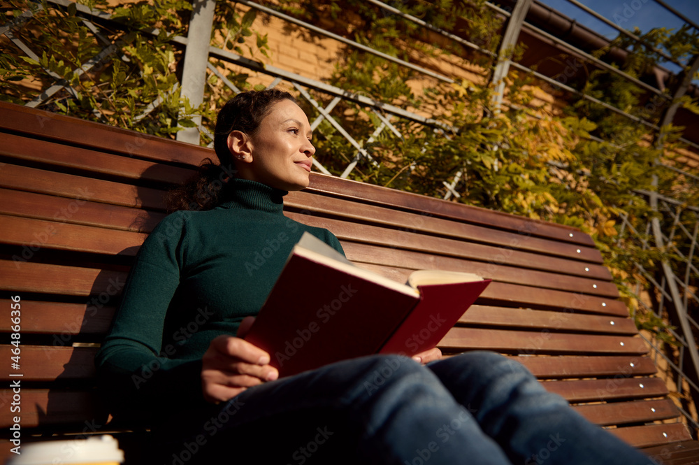 Fototapeta premium Relaxed pretty woman in casual denim and green pullover reading a book sitting on a wooden bench enjoying a warm sunny autumn weekend in country park, away from the hustle and bustle of the city