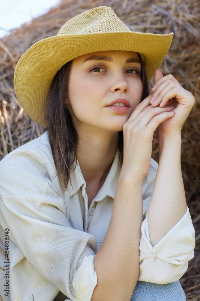 Young brunette woman in a white shirt and jeans is sitting on the rolls of hay.