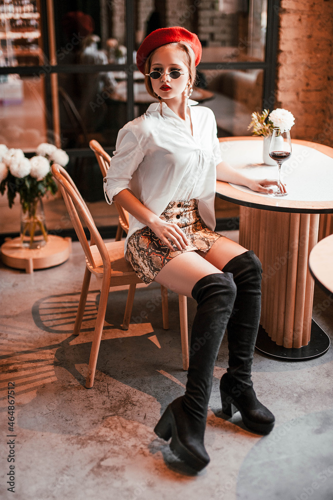 Obraz premium blonde girl with red lips in a white blouse in a snake skirt in sunglasses in a red beret with a glass of wine in a restaurant in a relaxing atmosphere