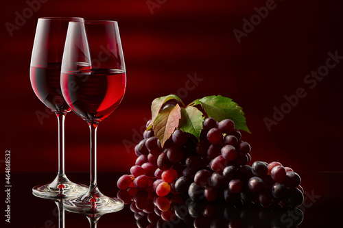 Two glasses of red wine and a bunch of grapes on a glossy table.