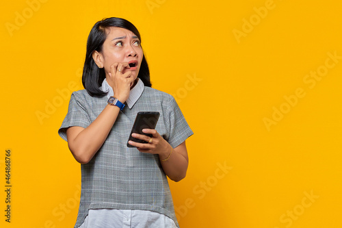 Young Asian woman holding smartphone looks anxious and worried afraid of something isolated over yellow background
