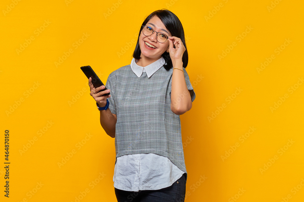 Portrait of Cheerful young Asian woman holding smartphone and eyewear looking at camera over yellow background Portrait of Cheerful young Asian woman holding smartphone and eyewear looking at camera over yellow background