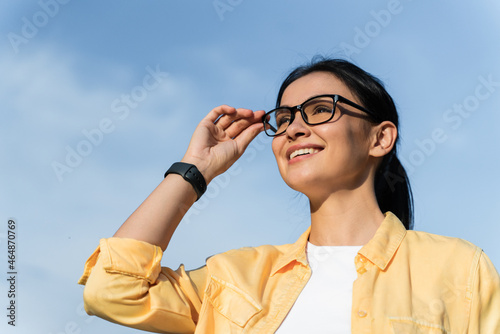 Looking at my perspectives. Low angle view of the satisfied woman touching her glasses and looking at the distance while walking at the street during the sunny day. Stock photo