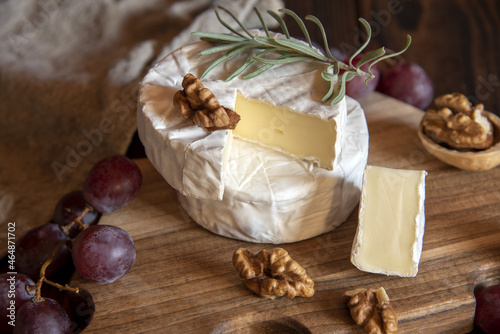 Cheese with white mold, nuts and grapes on a wooden cutting board.