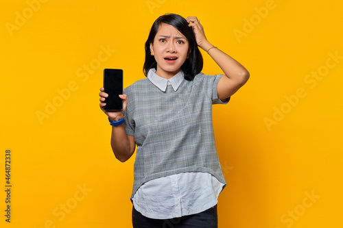 Portrait of beautiful asian woman holding smartphone touching her head and expressing concern on yellow background