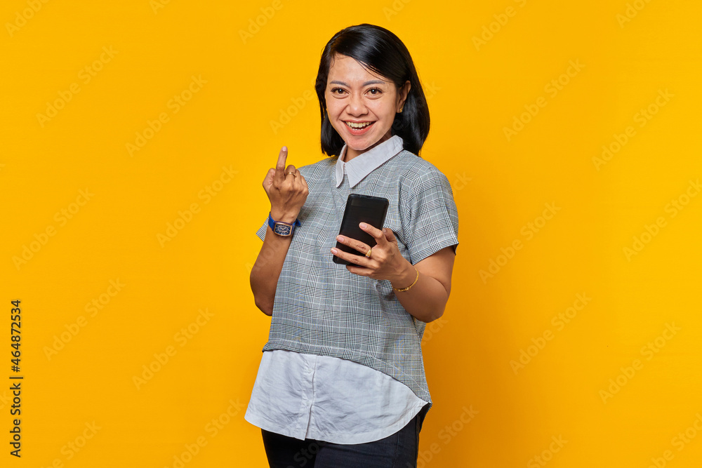 Portrait of cheerful asian woman pointing at camera and holding smartphone over yellow background Portrait of cheerful asian woman pointing at camera and holding smartphone over yellow background