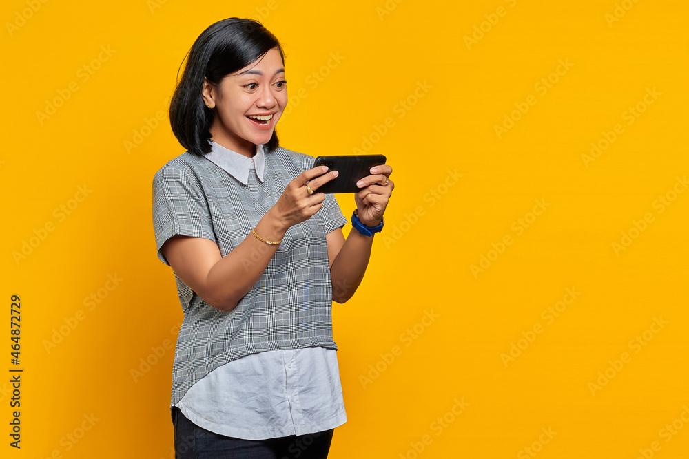 Portrait of cheerful young Asian woman playing video game on mobile phone over yellow background Portrait of cheerful young Asian woman playing video game on mobile phone over yellow background