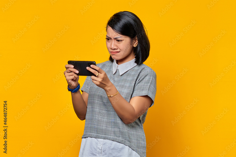 Portrait of angry young Asian woman playing video game on mobile phone over yellow background Portrait of angry young Asian woman playing video game on mobile phone over yellow background