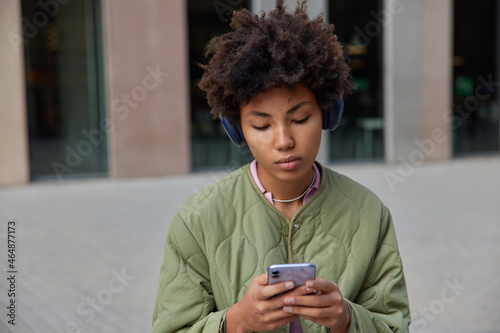 Charming young woman listens music podcast during leisure uses application for listening audio songs holds mobile phone searches internet websites connected to wireless internet sits outdoors