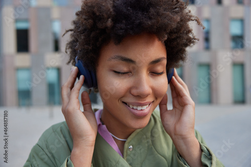 Close up shot of cheerful curly haired woman closes eyes from pleasure while listens favorite audio song via headphones wears jacket poses against blurred background enjoys music from playlist