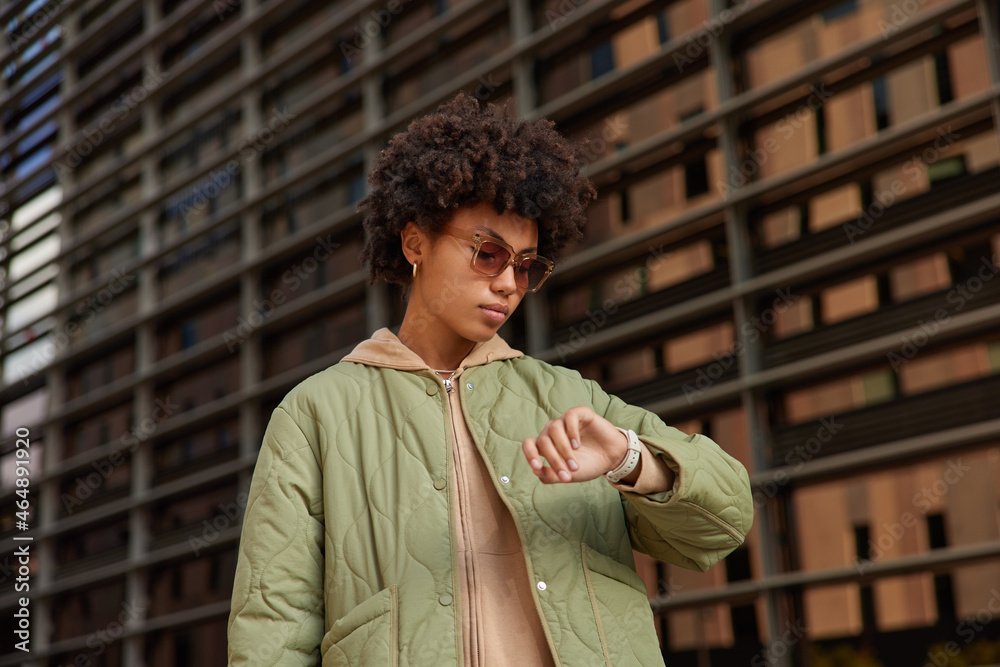 Ταπετσαρία Photo of serious curly woman hurries for work or meeting looks attentively at wrist watch checks time wears trendy sunglasses and jacket poses outdoors tracks clock