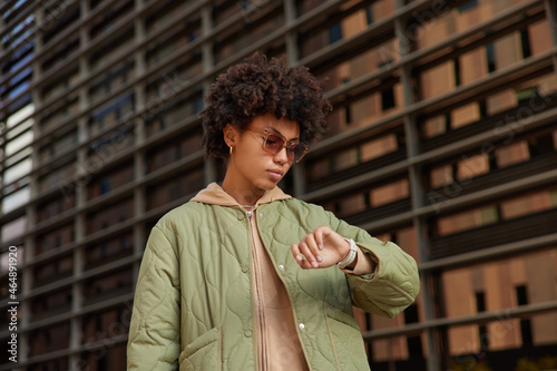 Bild auf Leinwand Photo of serious curly woman hurries for work or meeting looks attentively at wrist watch checks time wears trendy sunglasses and jacket poses outdoors tracks clock