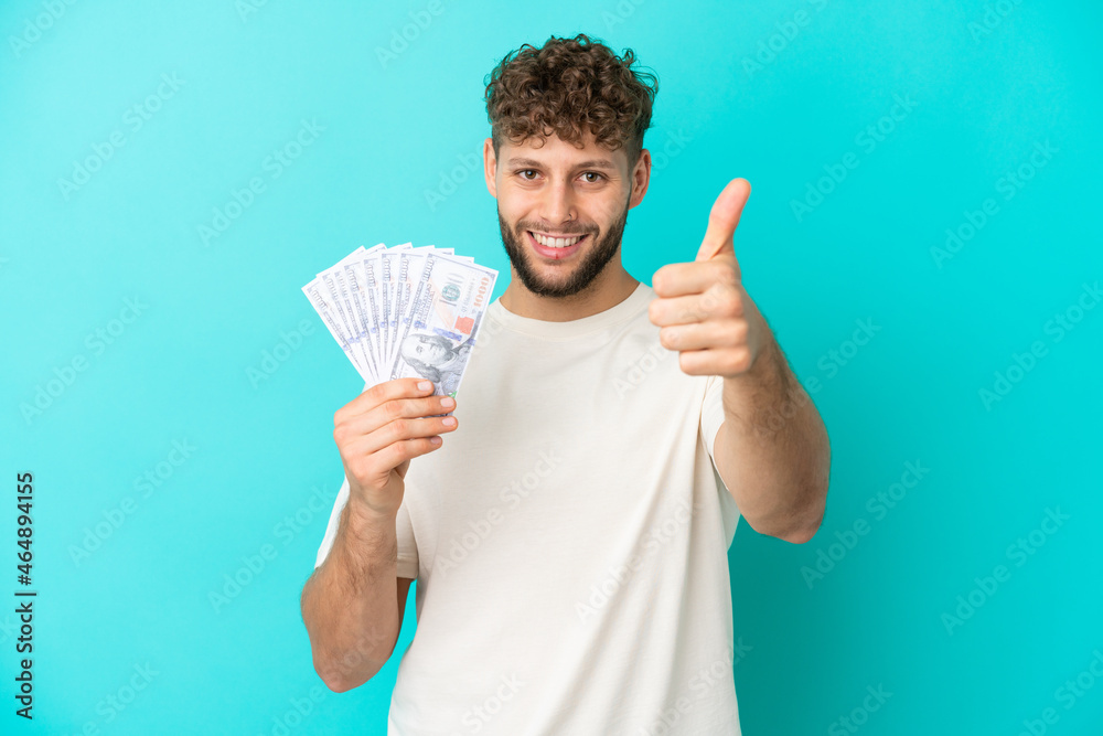 Young caucasian man taking a lot of money isolated on blue background with thumbs up because something good has happened Young caucasian man taking a lot of money isolated on blue background with thumbs up because something good has happened