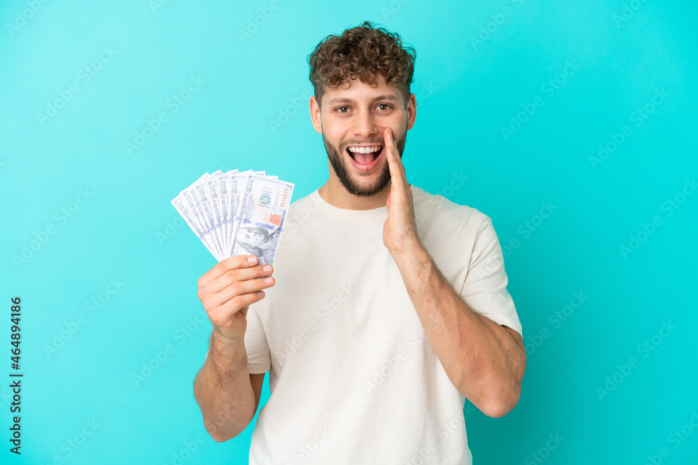 Young caucasian man taking a lot of money isolated on blue background with surprise and shocked facial expression Young caucasian man taking a lot of money isolated on blue background with surprise and shocked facial expression