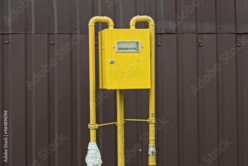 one gas meter in a yellow metal box with iron pipes outside by the brown wall of the fence