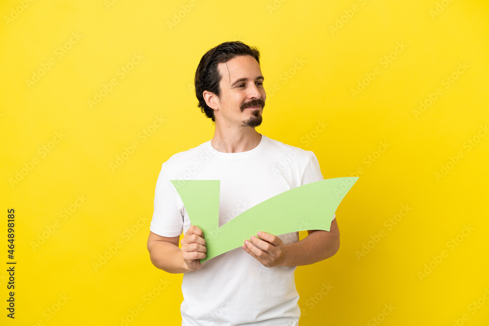 Young caucasian man isolated on yellow background holding a check icon and looking side Young caucasian man isolated on yellow background holding a check icon and looking side