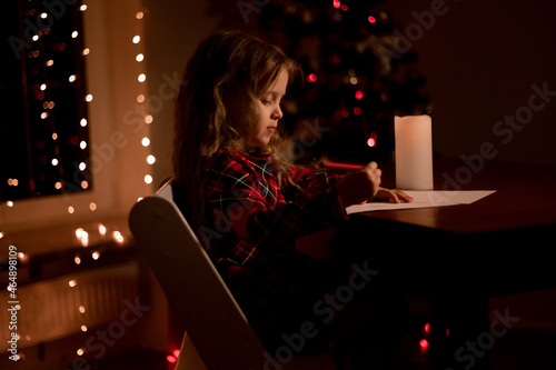 beautiful little preschool girl in the evening at home writes letter for santa claus, makes a wish and presents. Christmas tree, garlands, Christmas lights. Waiting for holiday, advent calendar