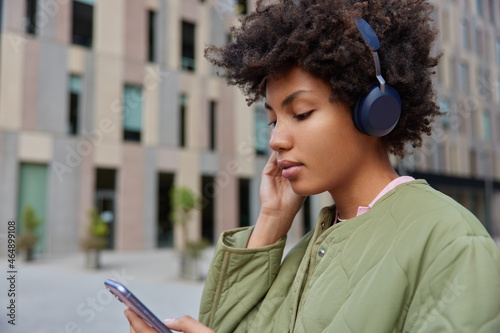 Sideways shot of pretty woman listens audio book via headphones installs media app on cellular wears jacket poses against blurred building background. Female lover of music strolls outdoors.