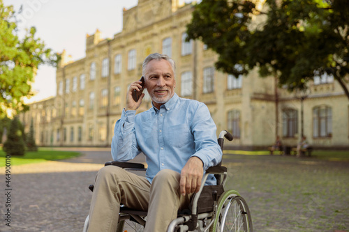 Smiling aged man recovering patient in wheelchair looking away while talking on the phone, spending time alone in the park near hospital