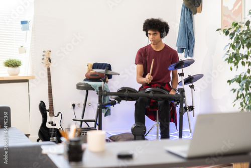 Billede på lærred Teenage guy with headphones hitting drumset while sitting in front of table with