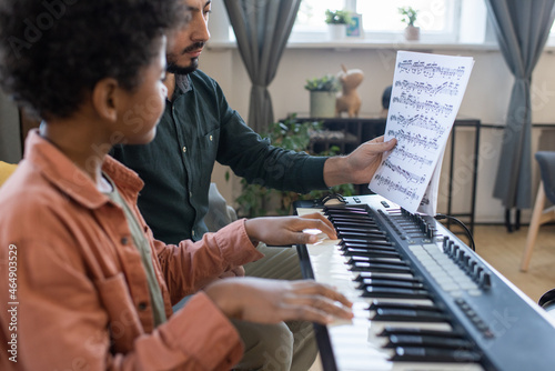 Teacher of music showing little schoolboy paper with notes while both sitting by piano keyboard at lesson