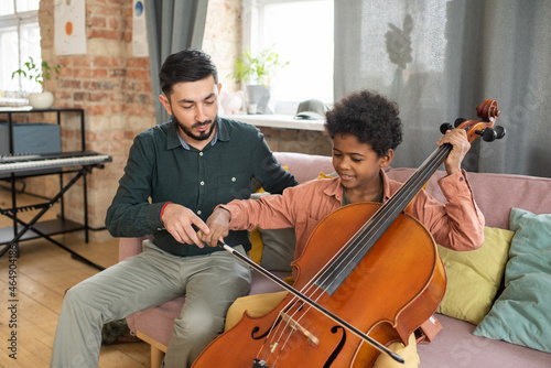 Cute biracial boy playing cello while sitting on couch next to his teacher durin Fototapet